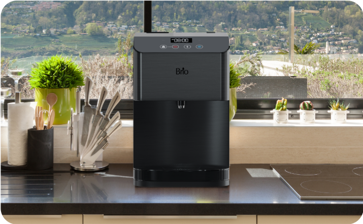 Brio black countertop water dispenser placed in a modern kitchen on a dark counter, with plants, knives, and utensils by the window overlooking a scenic outdoor view.